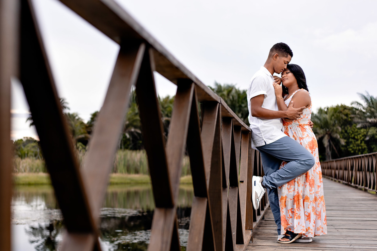 Momento íntimo de casal na ponte de madeira da Praia do Forte — noivo e noiva com testas unidas em pose romântica com lago ao fundo, ensaio pré-casamento por Filipe Arreciguelli