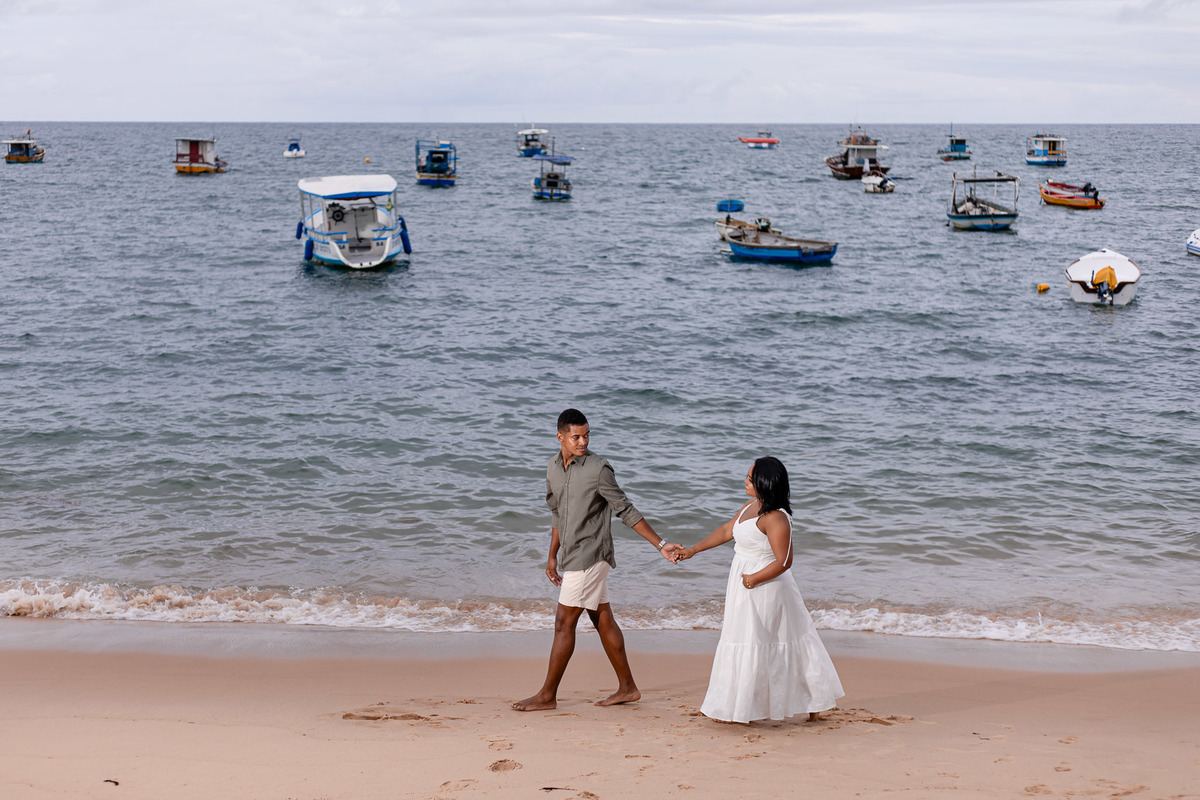 ensaio pré-casamento praia Bahia barcos, fotografia casal praia Salvador, ensaio pré-nupcial Praia do Forte barcos, fotógrafo casamento praia do forte bahia, casal mãos dadas praia fotografia, vestido branco ensaio praia noiva, fotografia litoral baiano e