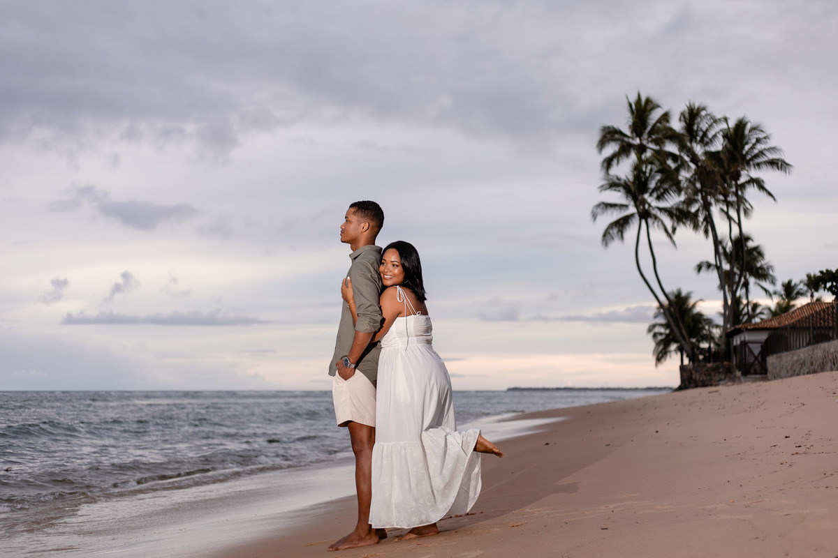 Casal em ensaio pré-casamento na Praia do Forte — noiva com vestido branco abraçada nas costas do noivo ao entardecer com coqueiros e mar ao fundo, fotografia por Filipe Arreciguelli