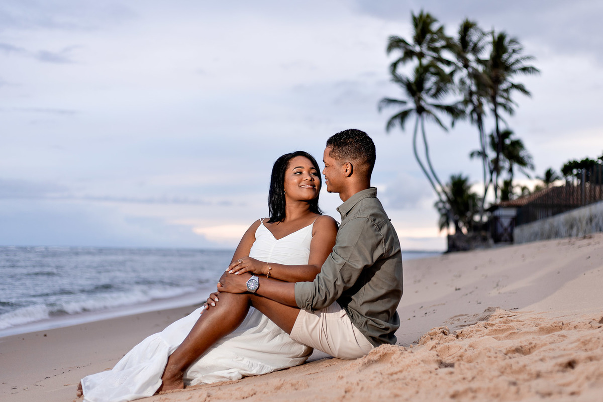 ensaio pré-casamento luz entardecer Praia do Forte, fotografia casal sentado luz dourada Salvador, ensaio pré-nupcial golden hour praia Bahia, fotografo casamento luz natural entardecer Bahia, casal sentado areia luz suave fotografia, ensaio noivos expres