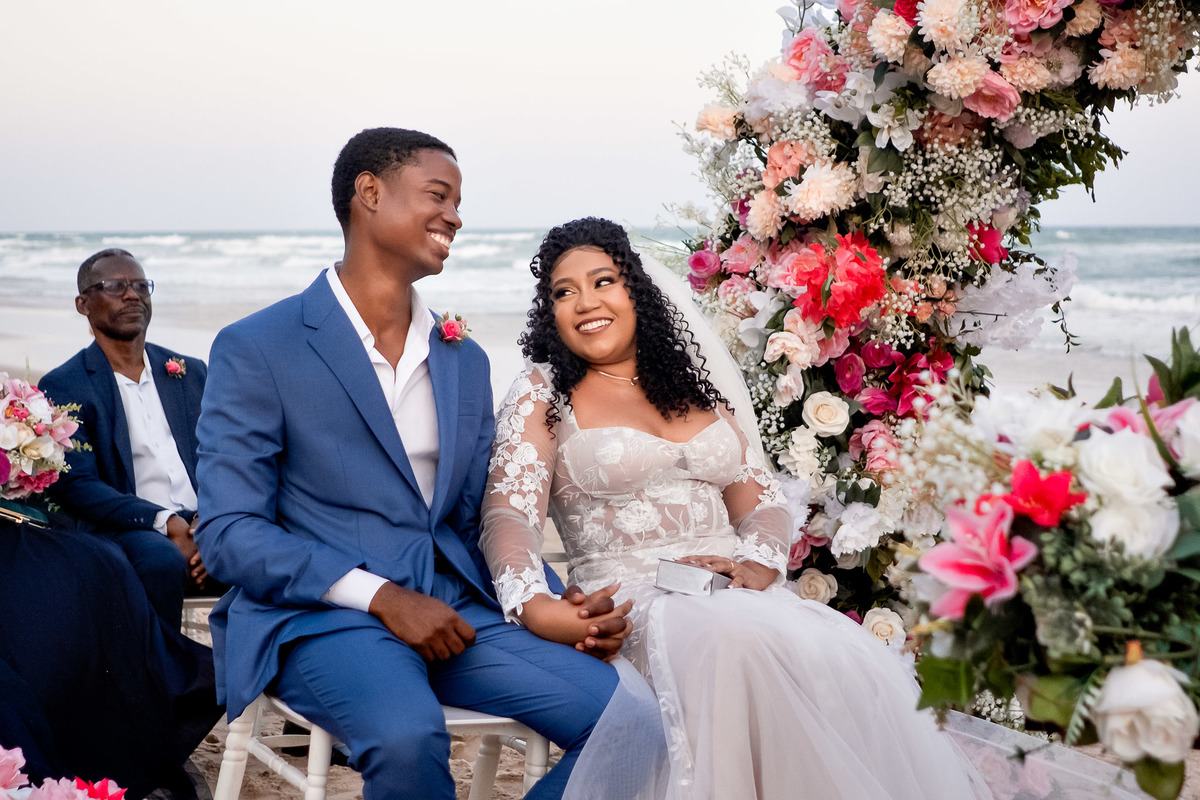 Noivos sorrindo sentados sob arco de flores durante casamento na praia em Salvador