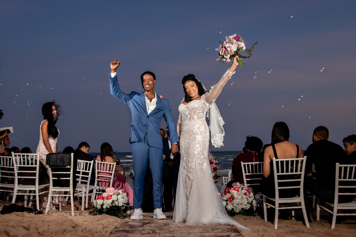 Casal comemorando com convidados ao fundo durante casamento na praia em Salvador