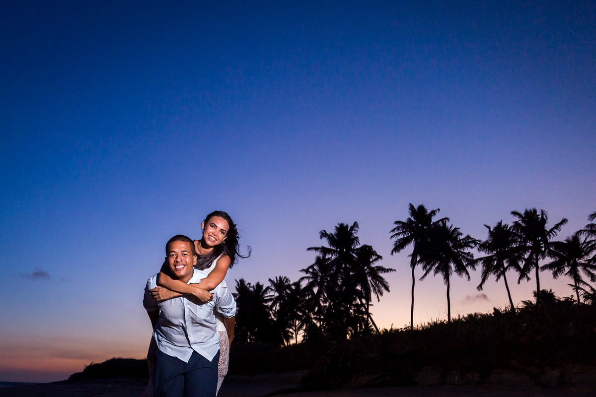 casal correndo pela praia com noiva nas costas do noivo com o por do sol no fundo embelezando a imagem