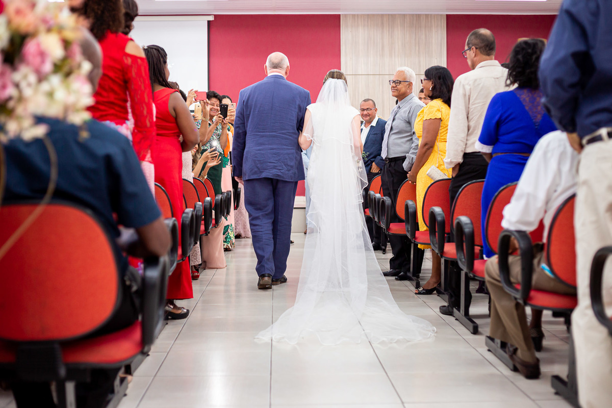 fotografia tirada na hora da entrada da noiva com o pai de costas mostrando todo o véu da noiva e o vestido durante a cerimônia em um casamento de dia