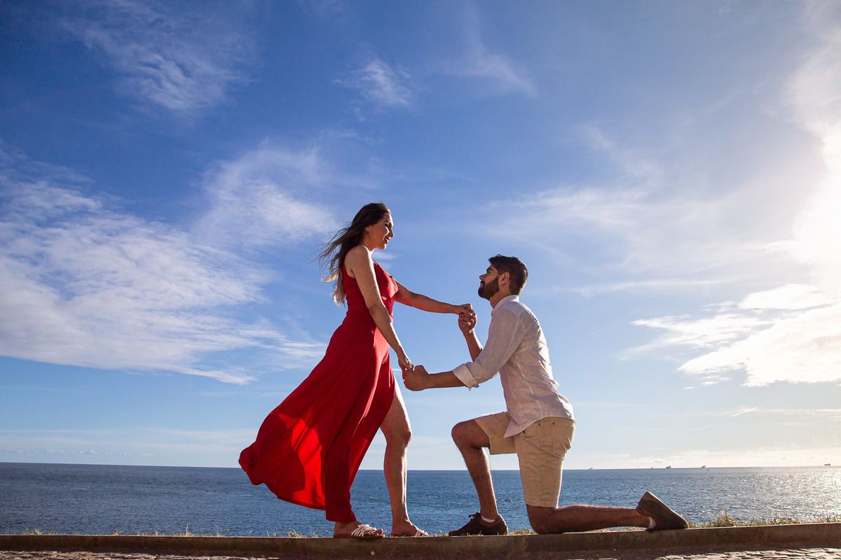 Foto de noivo ajoelhado segurando a mão da noiva de vestido vermelho longo na praia