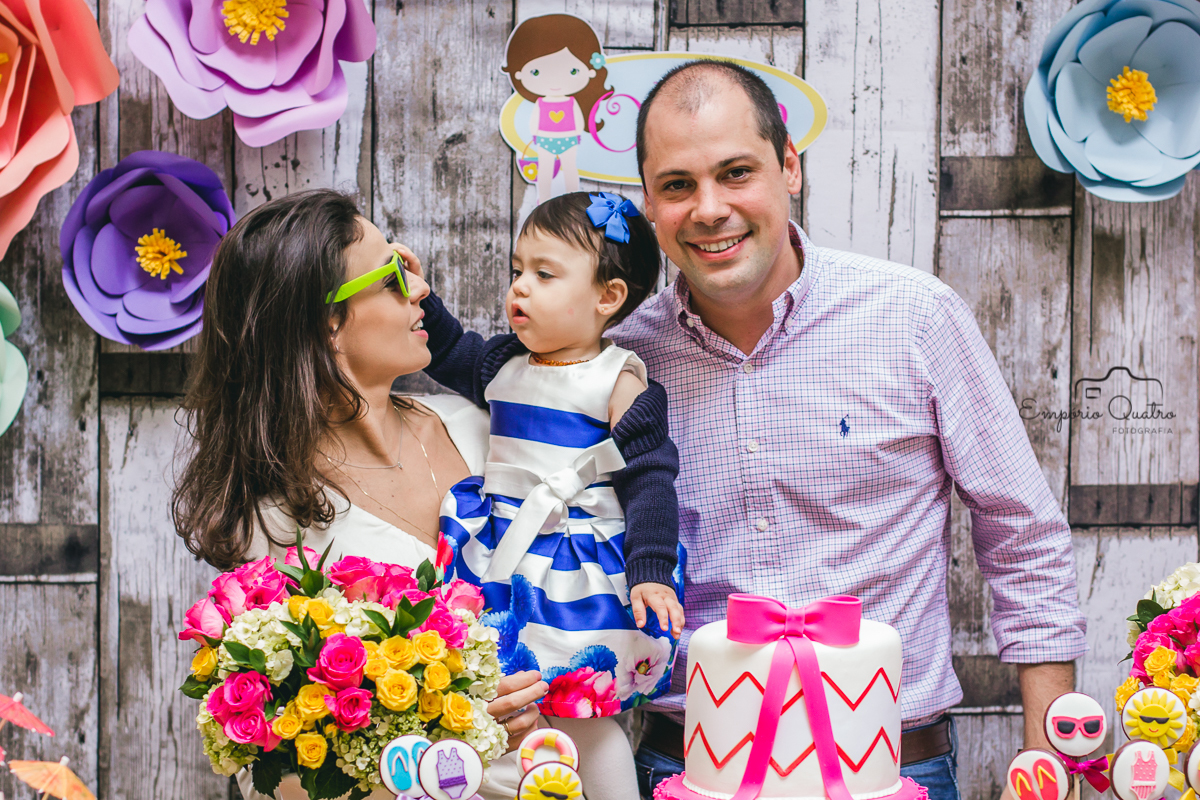 fotografia aniversário aniversariante no colo de seus pais brincando com o óculos de sol da mamãe na mesa do bolo decorada no tema praia