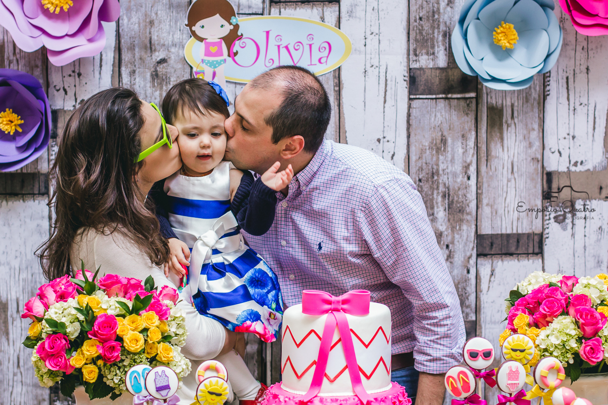 fotografia aniversário aniversariante no colo de seus pais sendo beijada no rosto na mesa do bolo decorada no tema praia