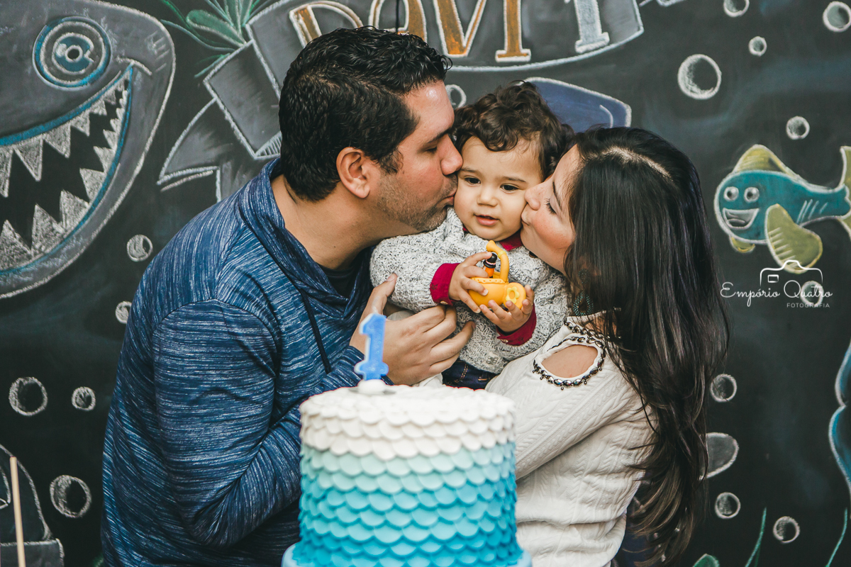 Aniversariante com seus pais na mesa do bolo brincando com a decoração Mundo Bita enquanto seus pais o beijam