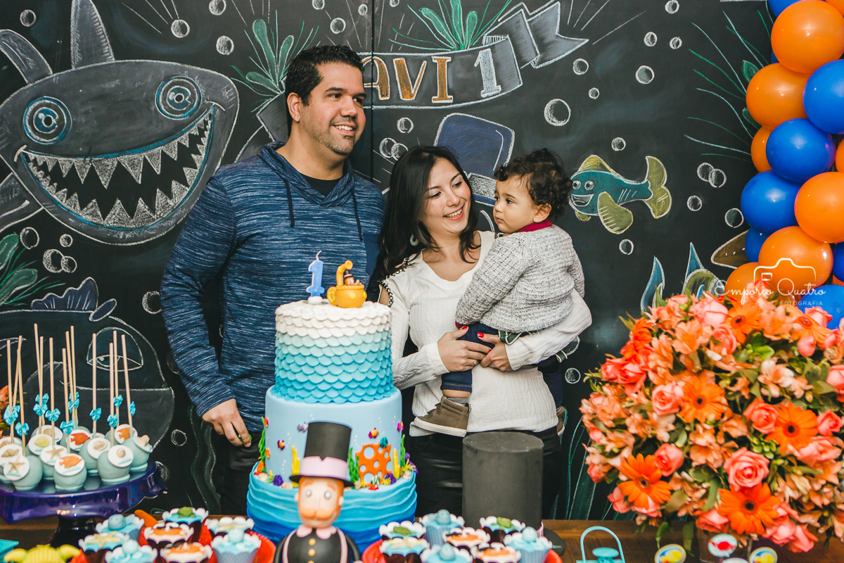 Aniversariante no colo da mãe ao lado de seu pai na mesa do bolo com decoração Mundo Bita