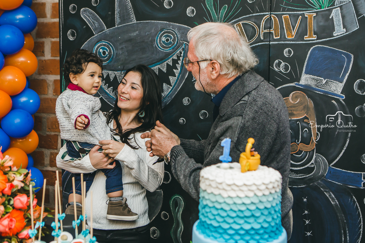 Aniversariante no colo de sua mãe ao lado de seu avô na mesa de bolo com decoração Mundo Bita