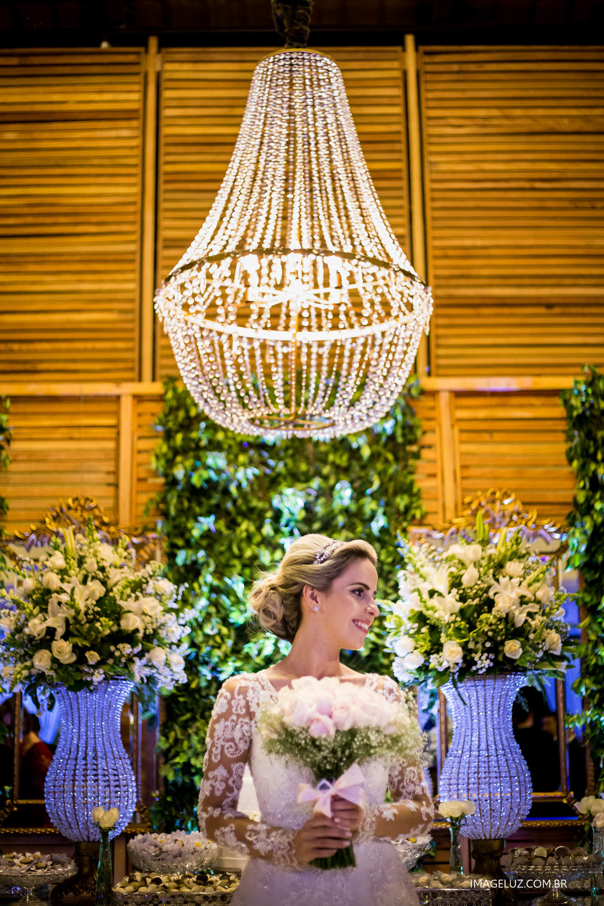 Noiva em frente a mesa de bolo, com um lustre lindo de fundo.