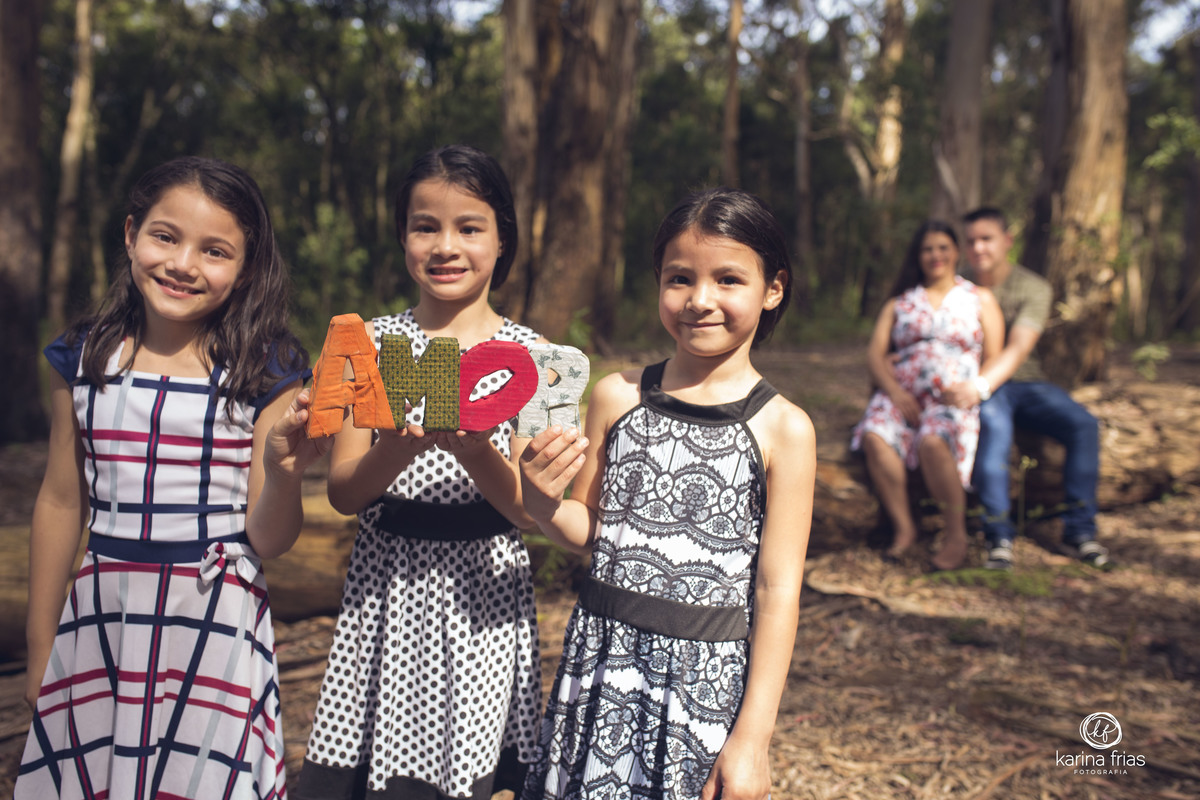 OS PAIS FICAM DESFOCADOS AO FUNDO ENQUANTO AS MENINAS SEGURAM A PLACA AMOR NA FRENTE
