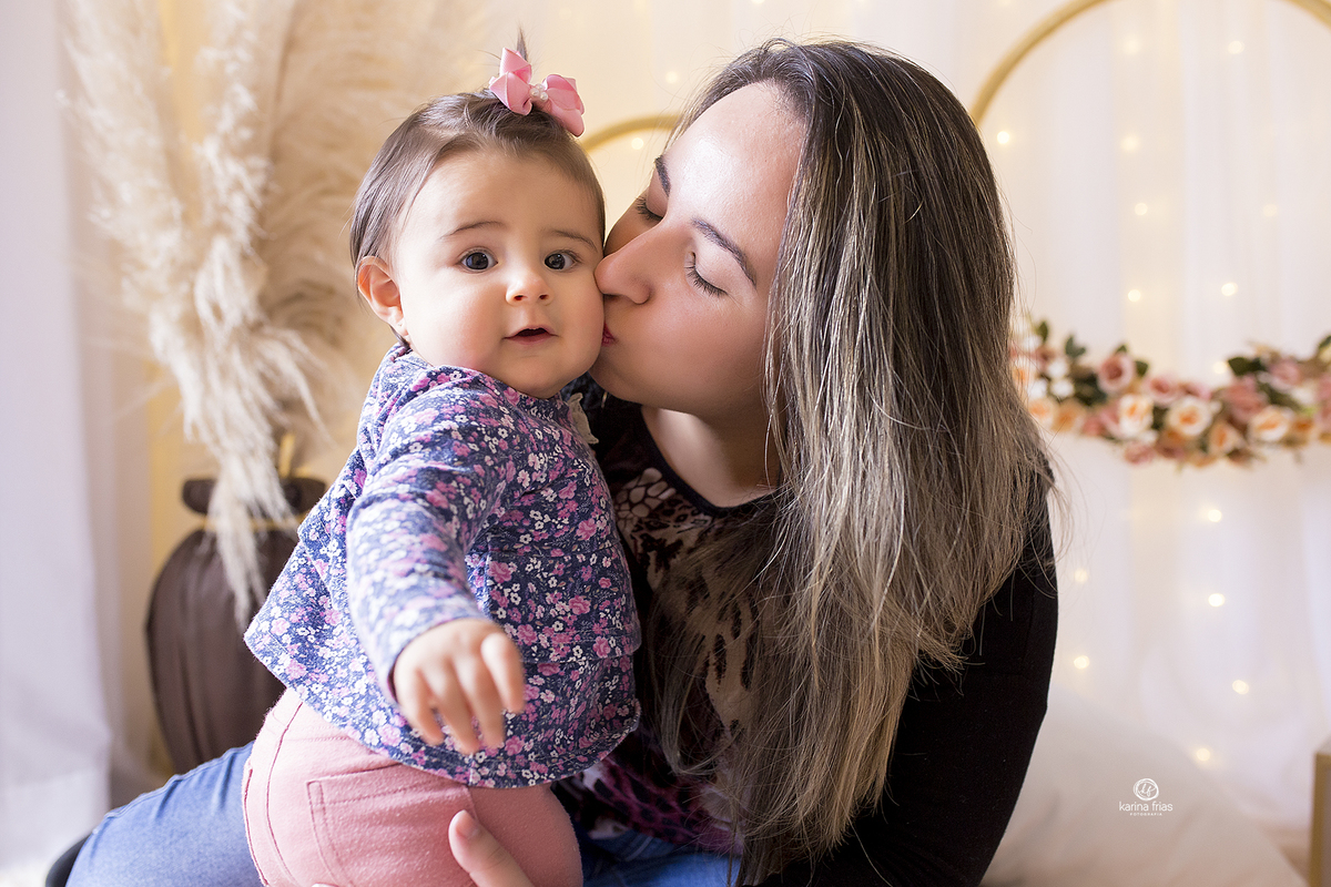 a menina olha para a fotografa de bebes de caxias do sul, enquanto recebe um beijo da mae