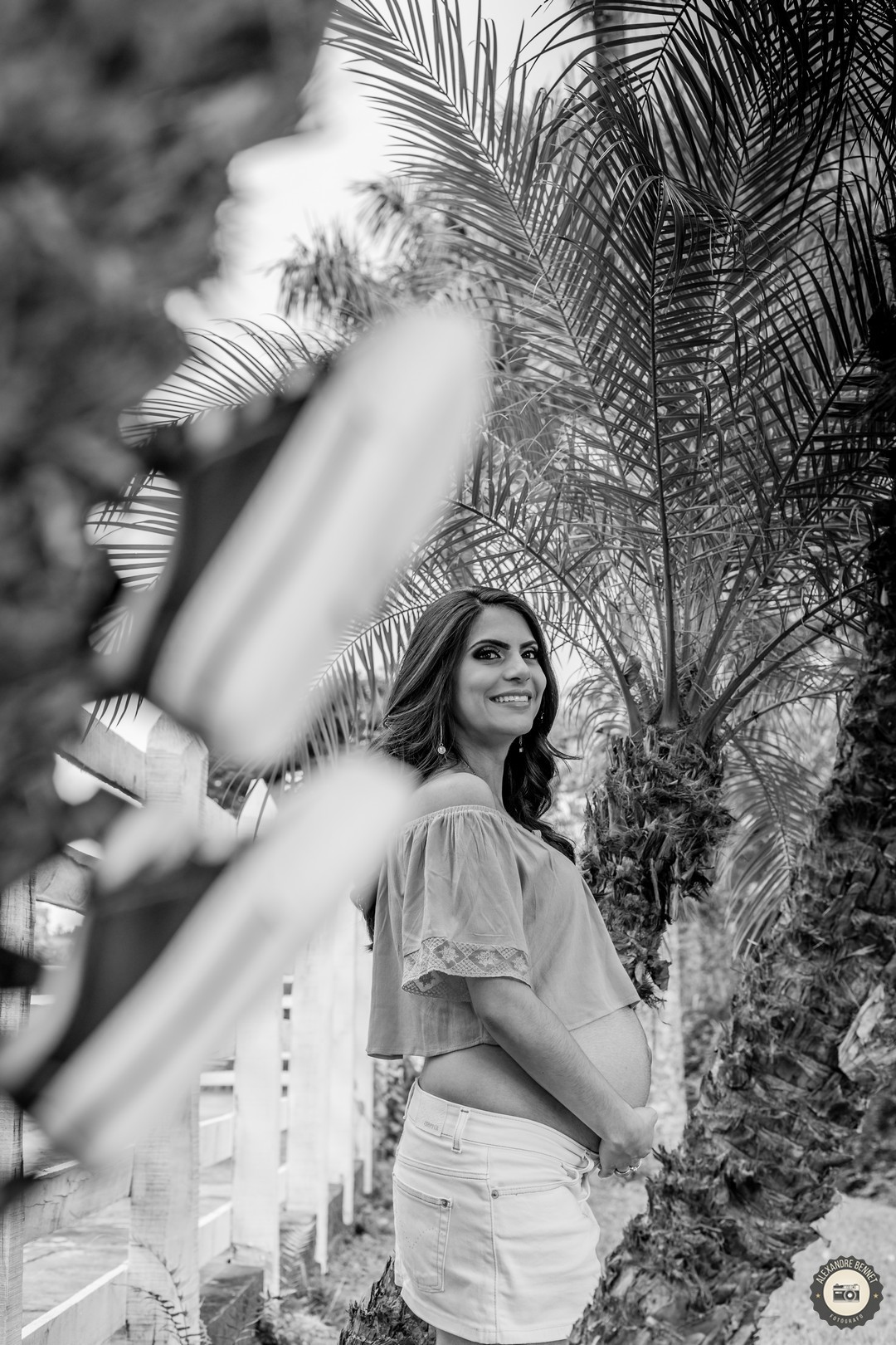 Com os sapatinhos do neném em primeiro plano a gestante aparece focada ao fundo sorrindo com as mãos na barriga 