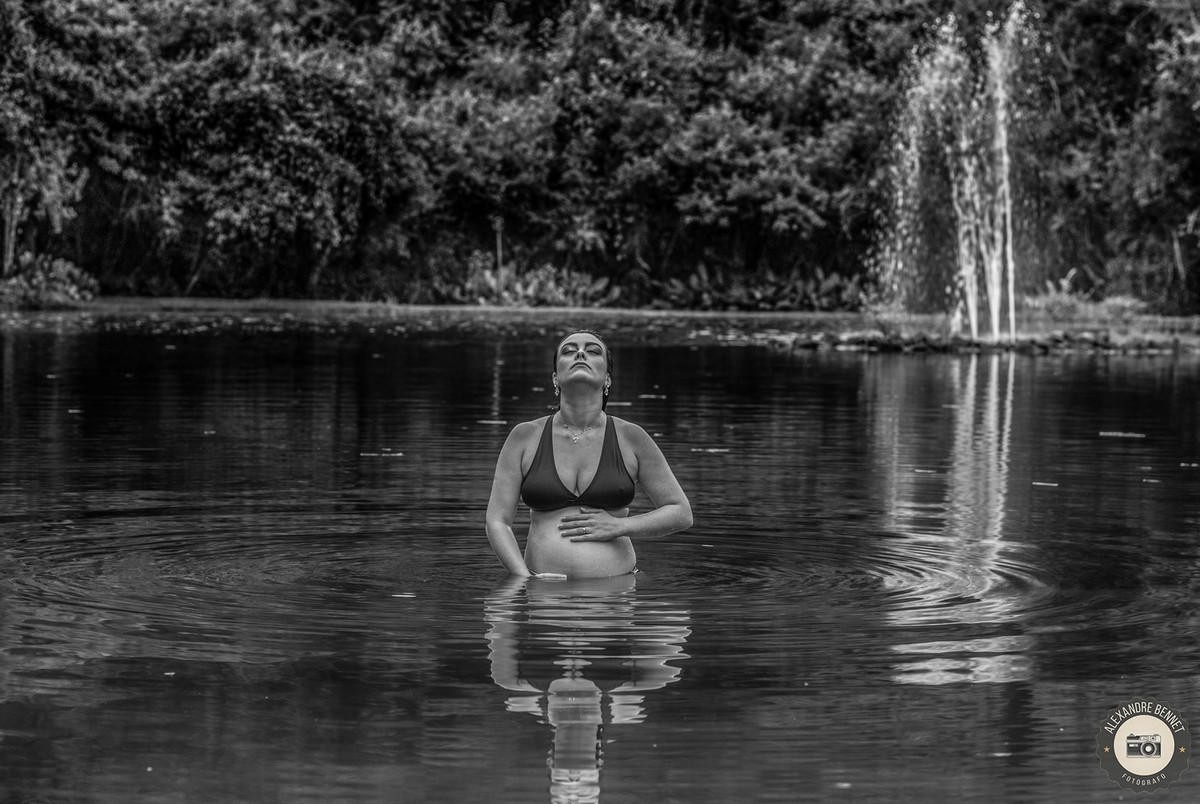 Dentro da lagoa com uma bela paisagem ao fundo a gestante fica de olhos fechados sentindo seu bebê