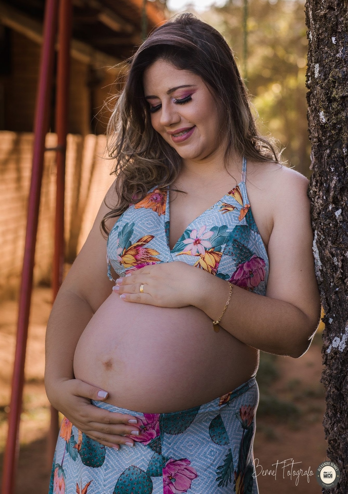 Gestante se encosta na arvore e sorri docimente enquanto olha para sua barriga 