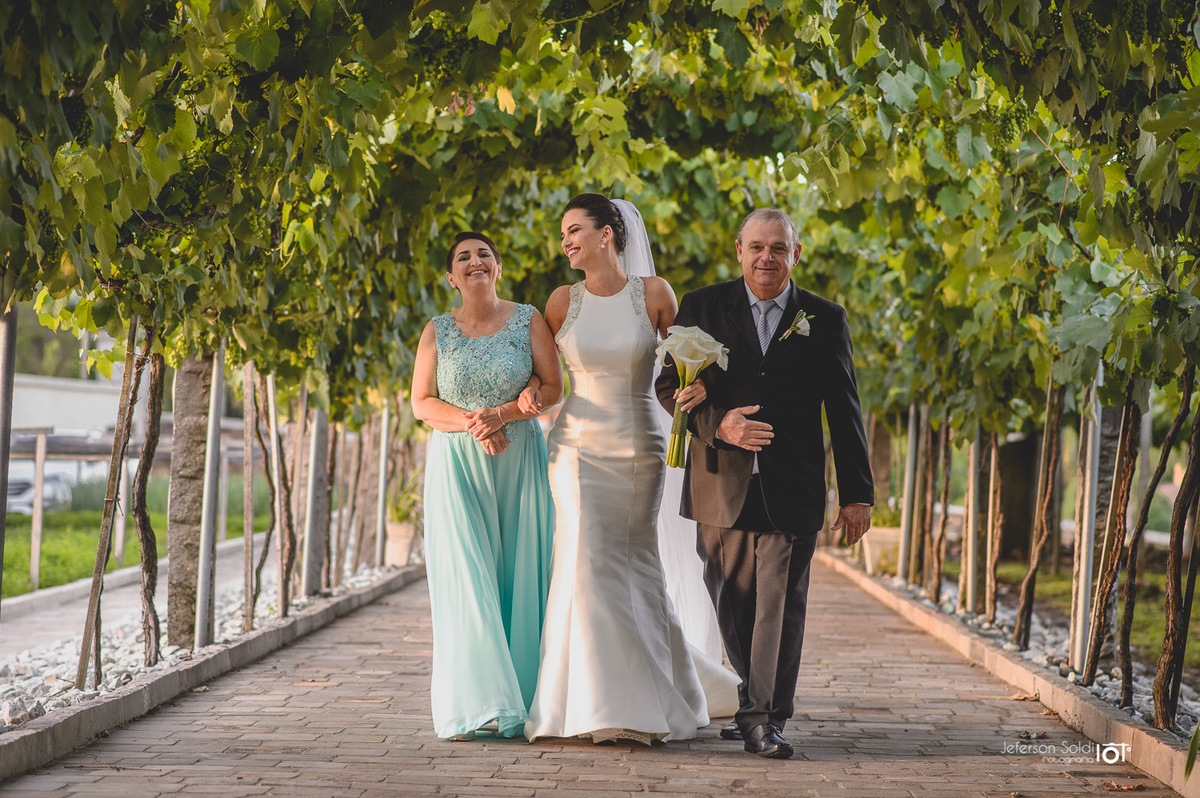 Noiva entrando no local da cerimônia acompanhada de seus pais , lado a lado.
