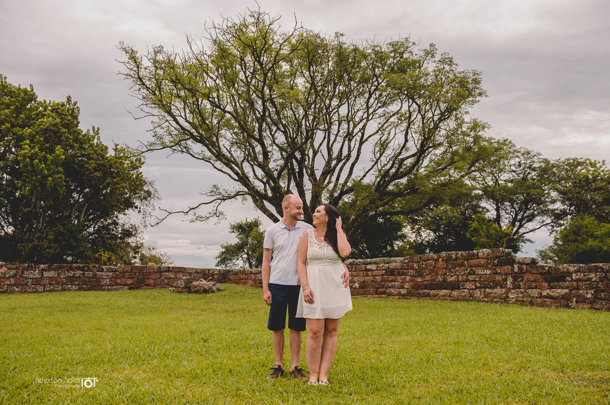 Casal se olhando em meio a grama e com uma figueira ao fundo. 