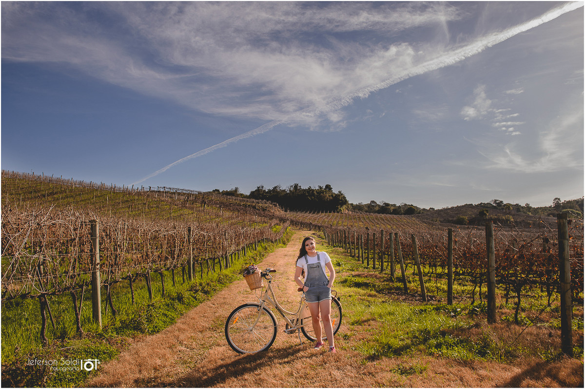 Julia parada ao lado de sua bicicleta em meio aos parreirais ao fundo um céu azul