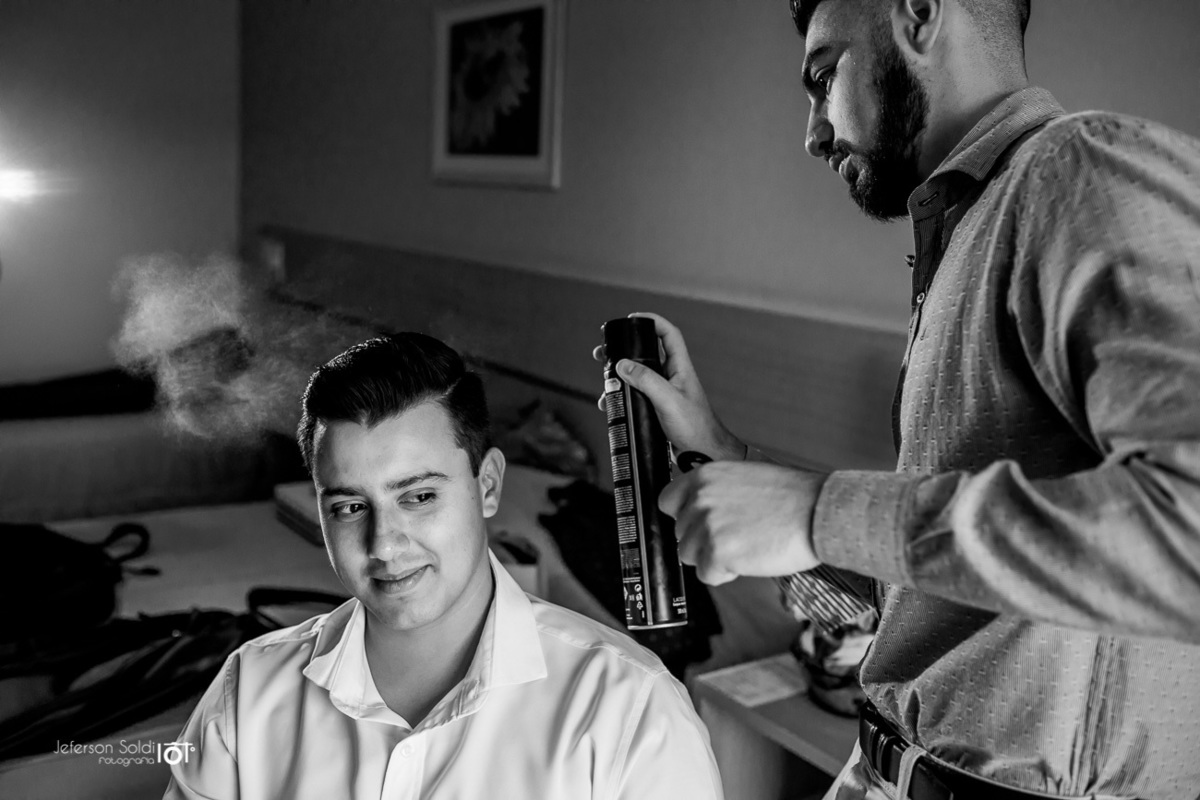 Cabelereiro passando spray no cabelo do noivo em uma foto preto e branco.