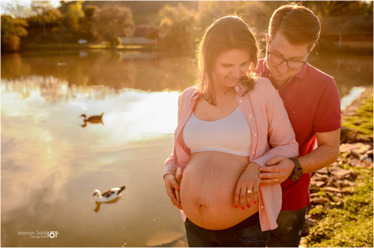 Casal abraçado em frente ao lago com dois patinhos ao fundo olhando para a barriga