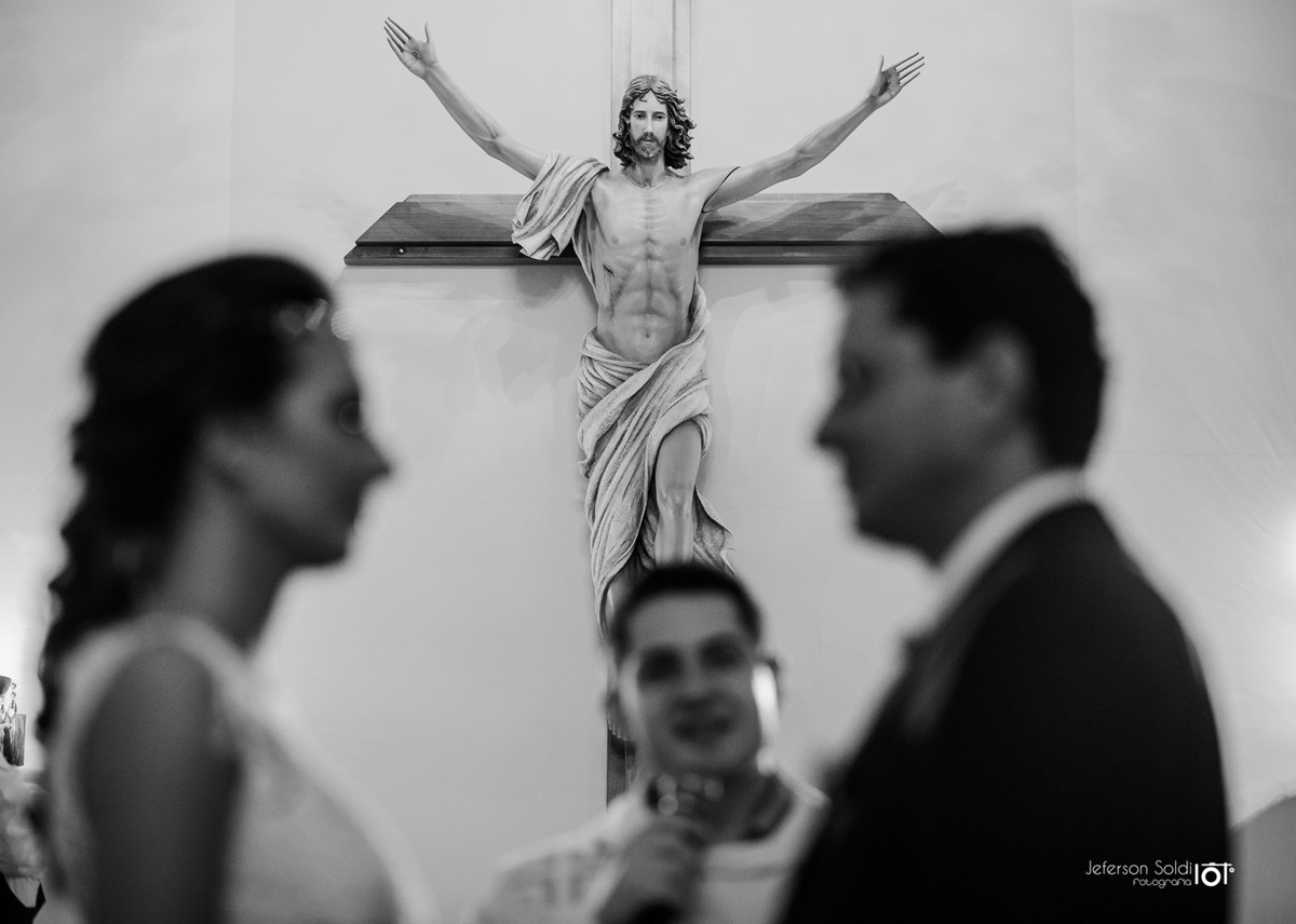 Noivos recebendo a benção do padre  sob a imagem de cristo na cruz ao alto.