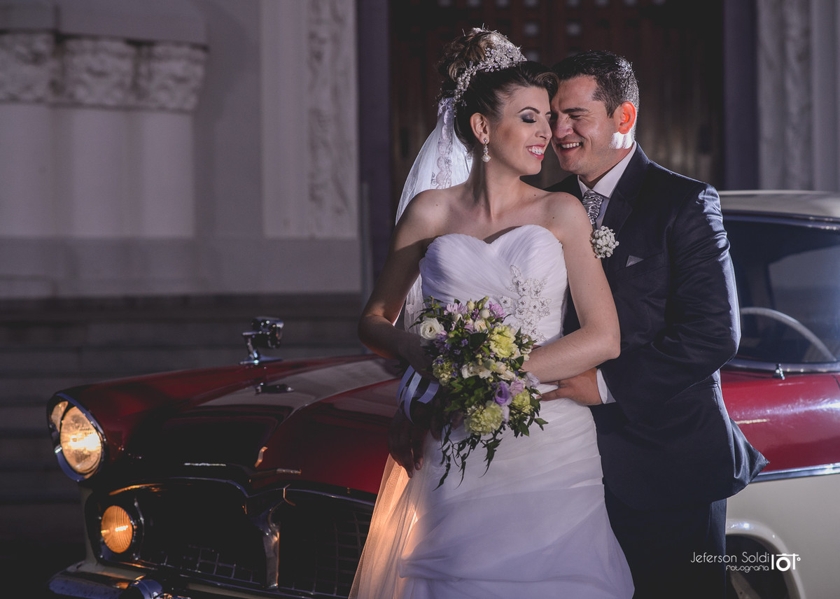 Noivos abraçados em frente a igreja, encostados no carro antigo.
