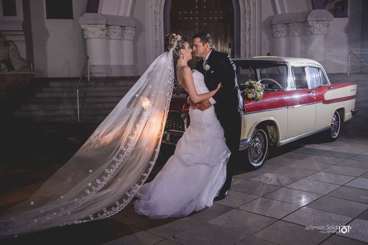 Noivos abraçados em frente a igreja e ao fundo o carro antigo que conduziu a noiva até a igreja.