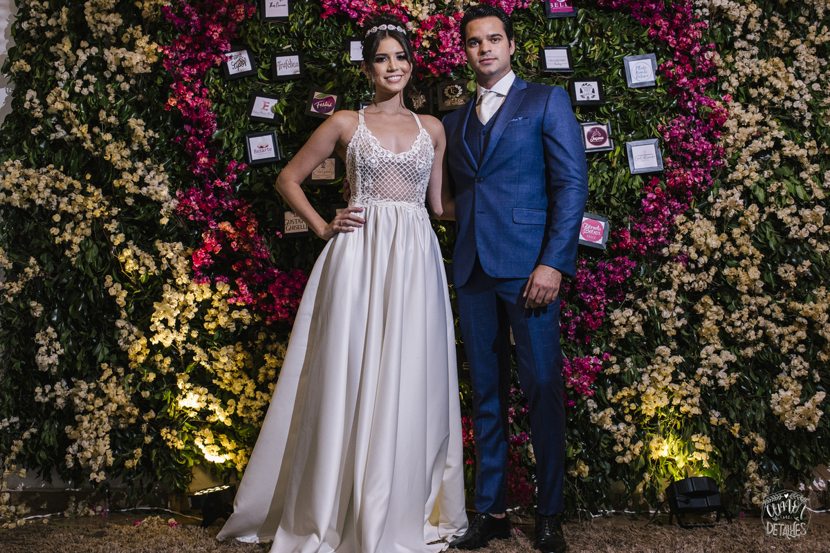 mulher de vestido em frente a parede de rosas com seu marido