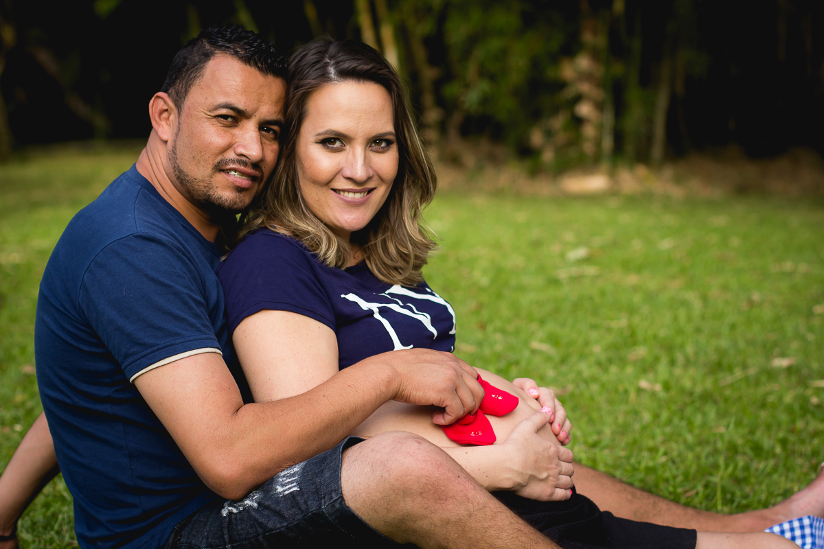 gestante sorridente com o seu marido sentados em um gramado e ele segura um par de sapatinho vermelho sobre a barriga dela