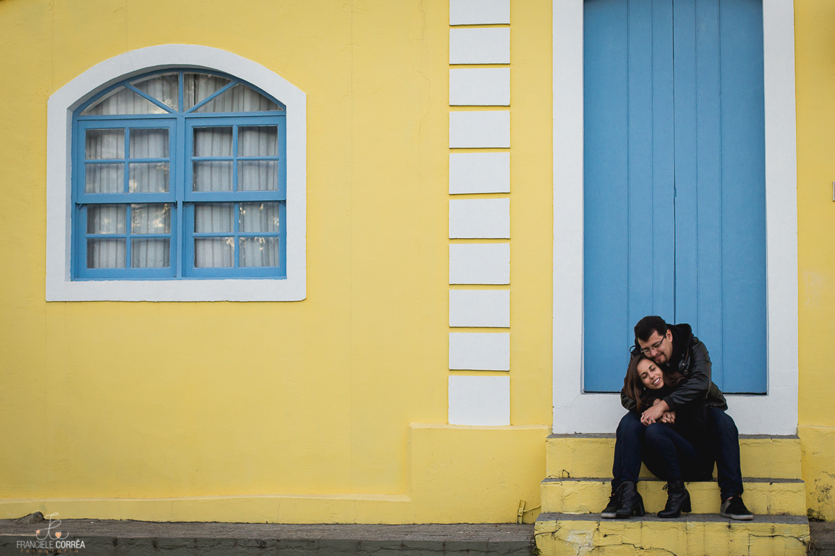 casal de noivos abraçados sentados em uma escada em frente a uma porta azul de uma casa amarela