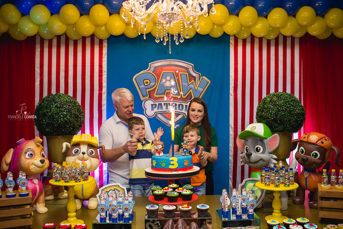 foto dos pais com os aniversariantes atrás do bolo batendo palma na hora do parabéns com a vela chafariz acesa