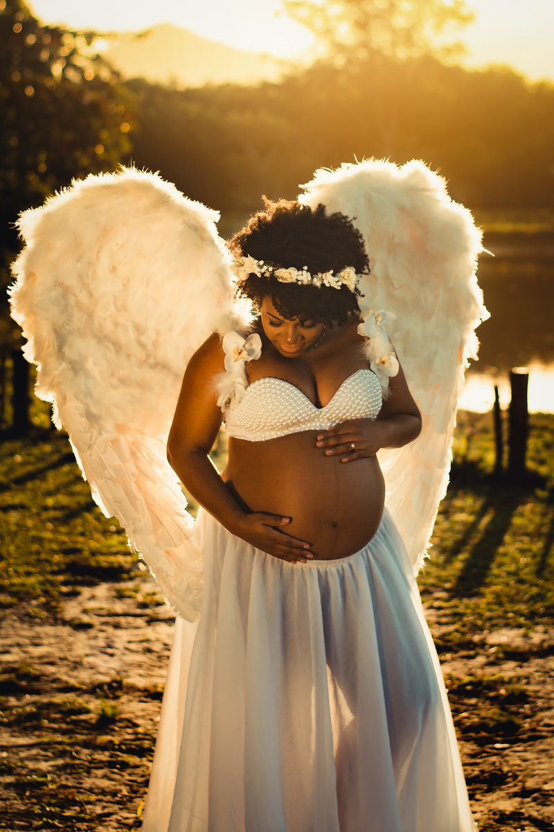 Mulher negra gestante usando asas de anjo na beira de um lago num belo final de tarde ao por do sol no Bosque da Barra
