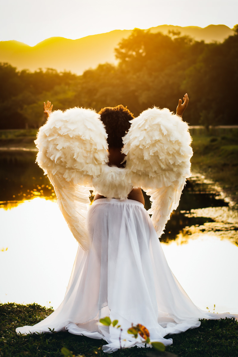Mulher negra gestante usando asas de anjo na beira de um lago num belo final de tarde ao por do sol no Bosque da Barra, agradecendo a Deus