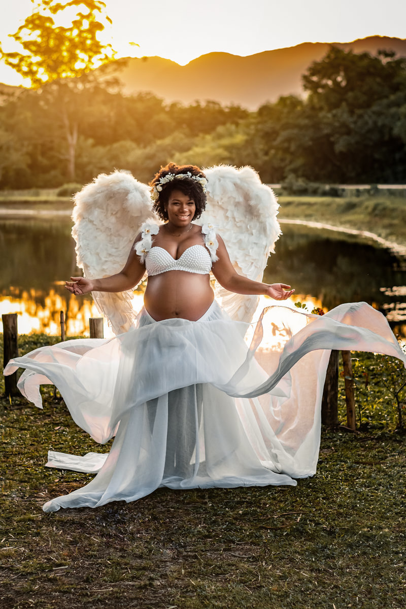 Mulher negra gestante com asas de anjo, na beira de um lago com tecidos da saia voando. Lago Bosque da Barra