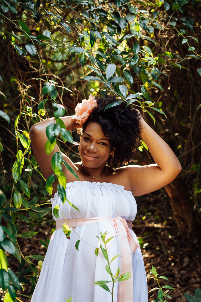 Mulher negra gestante com vestido branco em meio as folhas 
