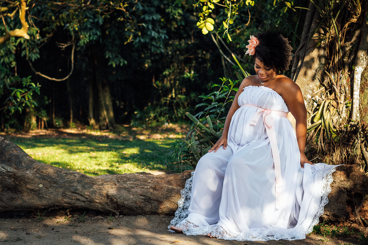 Mulher negra gestante usando vestido branco com uma fita rosa sentada em um tronco de arvore