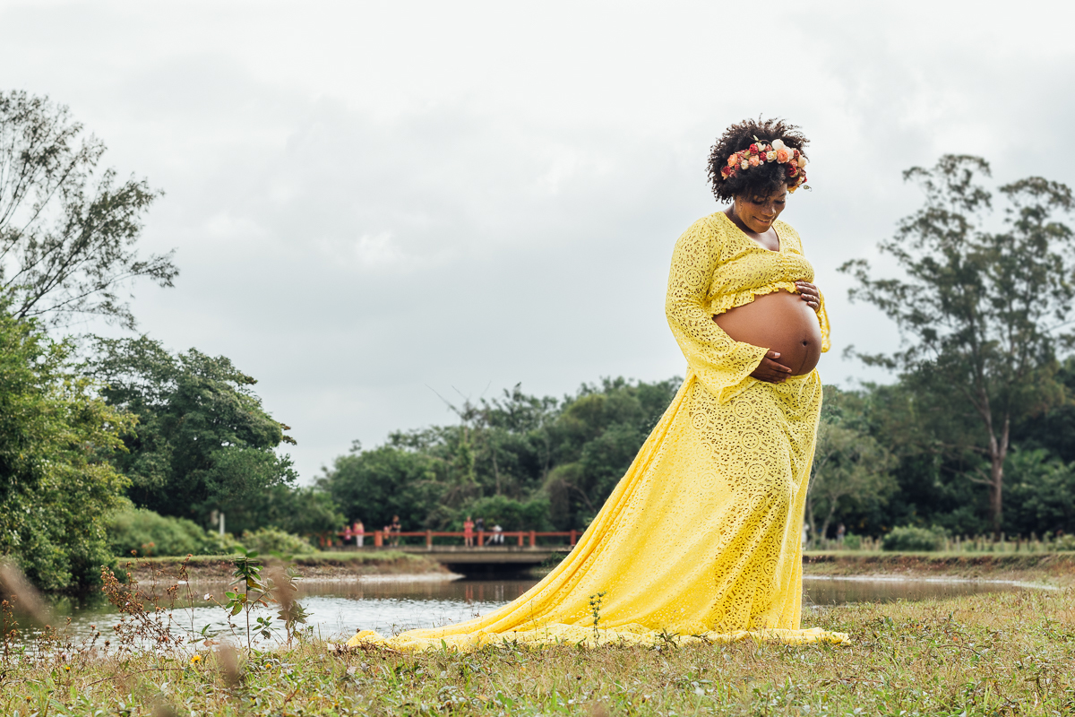 Mulher negra gestante usando vestido de renda amarelo e coroa de flores na beira do lago