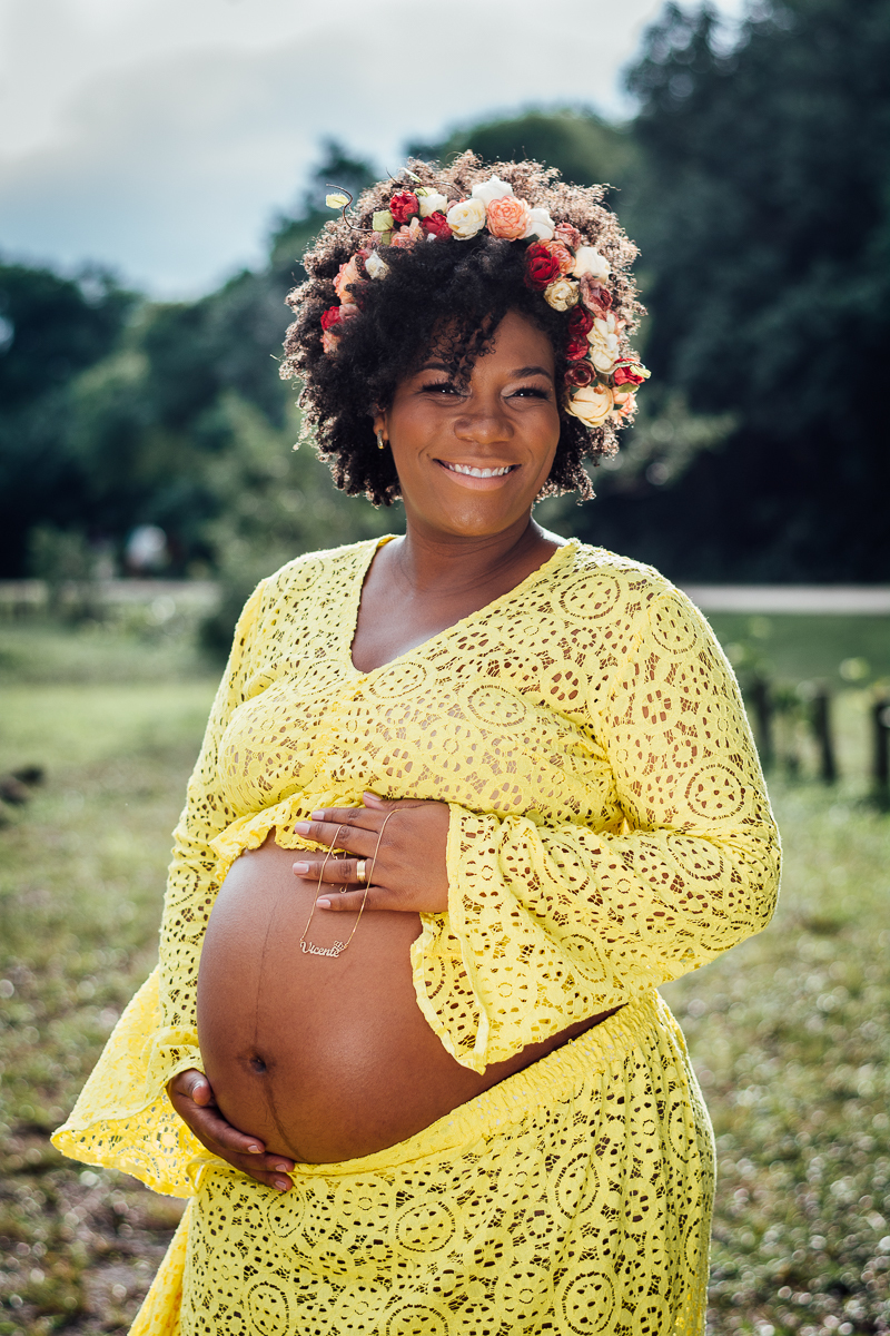 Mulher negra gestante usando vestido de renda amarelo e coroa de flores e segurando o cordão com nome do filho Vicente