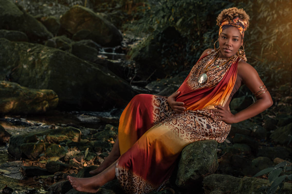 Gestante negra, com roupa estilo africana, usando argolas douradas e bracelete dourado de cobra. Esta sentada nas pedras em uma cachoeira
