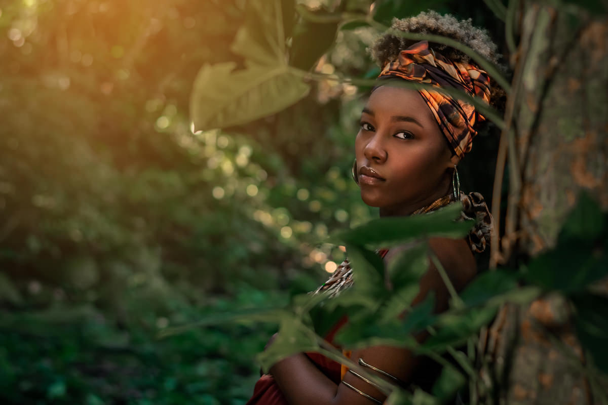 Gestante negra encostada na arvore, por tras de folhas verdes em meio a mata, vestindo uma roupa estilo africana, argolas douradas e bracelete dourado de cobra 