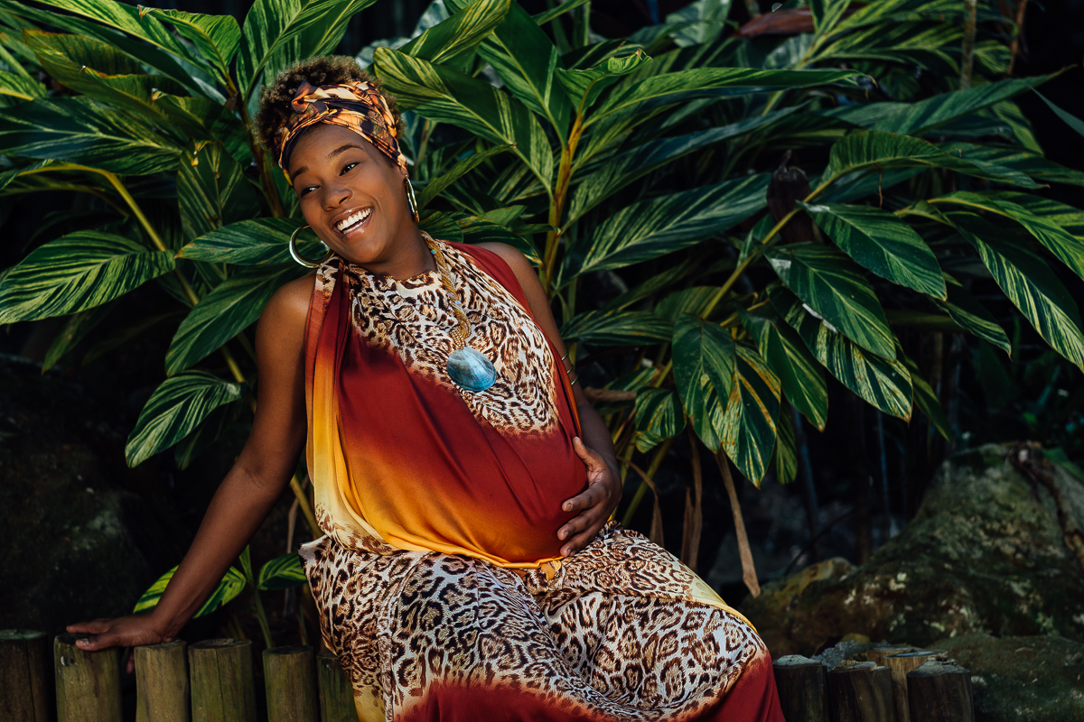 Gestante negra com vestido estilo africana, com belo sorriso, ao fundo folhas verdes, sentada no cercado, esta usando argolas douradas e faixa na cabeça