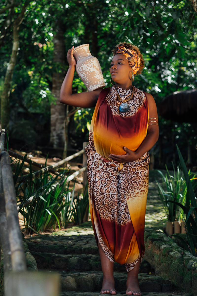 Gestante negra com vestido de estampa africana sentada na escada segurando um barro de jarro no ombro