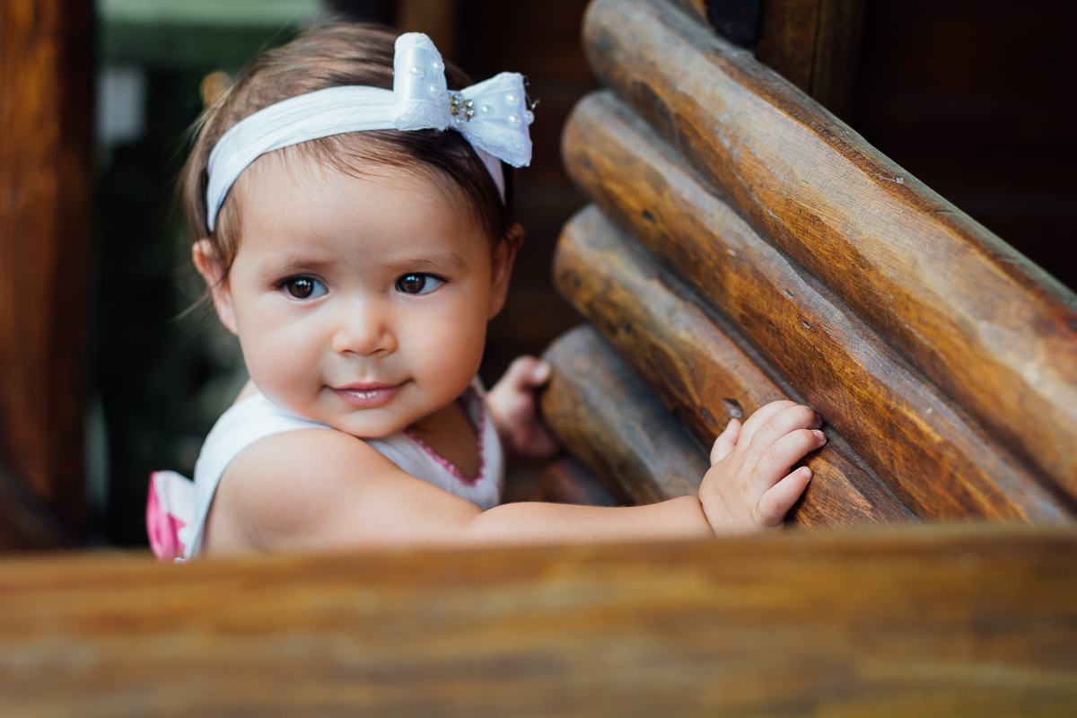 Bebê menina de vestido estampado e laço branco na cabeça brincando na casinha de madeira