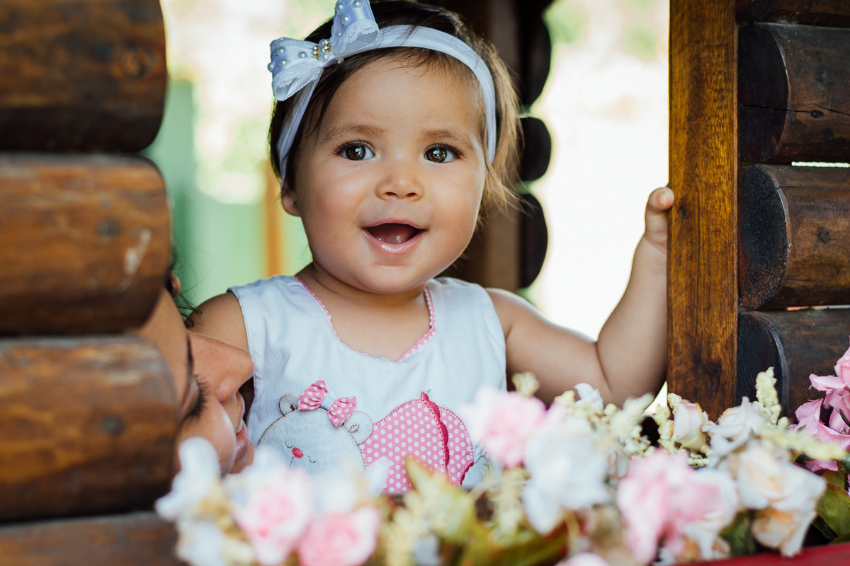 Bebê menina de vestido estampado e laço branco na cabeça brincando na janela com jardineira florida da casinha de madeira