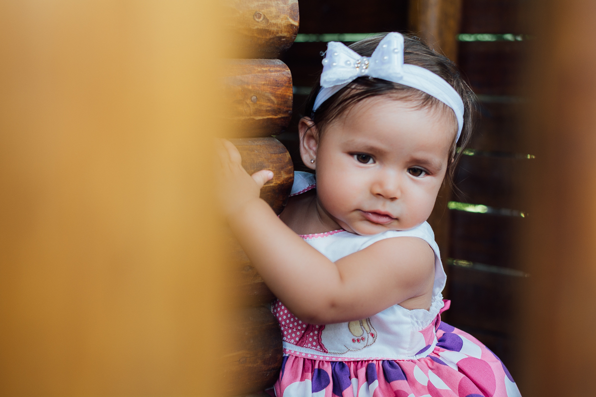 Bebê menina de vestido estampado e laço branco na cabeça brincando na casinha de madeira