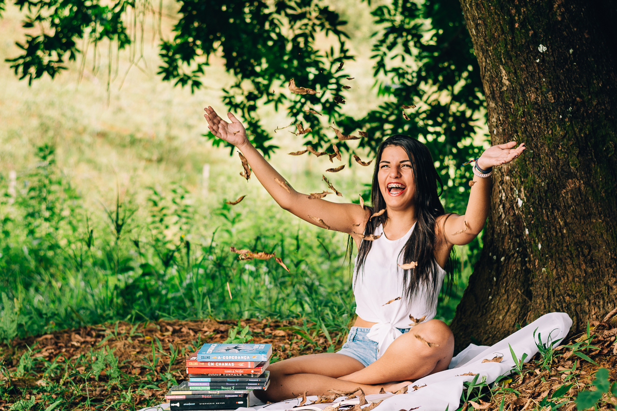 Menina com livros sentada de baixo da arvore e jogando folhas para o alto
