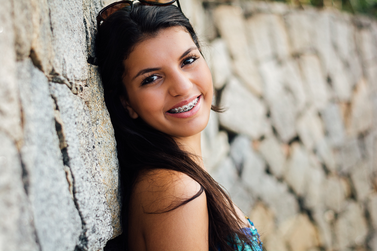 Encostada na parede de pedras sorrindo e de óculos escuros