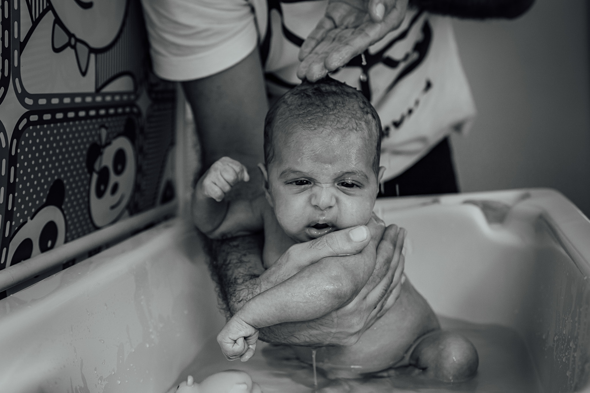 Pai dando banho no filho bebê, lavando sua cabecinha, enquanto ele faz uma cara de não estar muito feliz com o banho. 