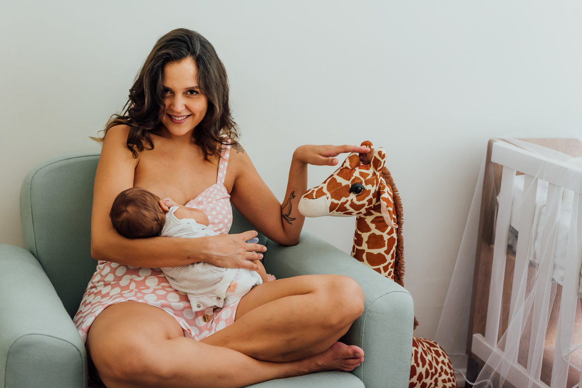 Mamãe sentada na cadeira ou sofá de amamentação, da mama para o bebê enquanto acaricia a girafa de pelúcia ao lado.